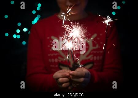 Weihnachtsfeier. Ein Kind hält ein brennendes Feuerwerk auf einem glänzenden Hintergrund mit leuchtend goldenen Weihnachtslichtern. Silvester. Dunkles Konzept. Stockfoto