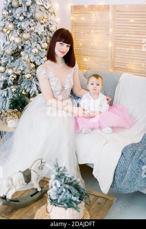 Hübsche Mutter mit ihrem Baby Mädchen, trägt in modischen eleganten Kleidern, sitzt auf stilvollen grauen Sofa auf dem Hintergrund von Weihnachtsbaum, Lichter und Stockfoto