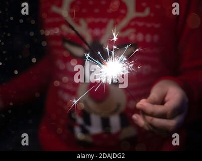 Weihnachtsfeier. Ein Kind hält ein brennendes Feuerwerk auf einem glänzenden Hintergrund mit leuchtend goldenen Weihnachtslichtern. Silvester. Dunkles Konzept. Stockfoto