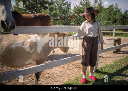 Lächelndes Mädchen in Cowboy-Kleidung streichelt kleines Pony im Paddock am sonnigen Sommertag. Betreuung der Tiere Konzept Stockfoto