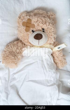 Kranker Teddybär mit Gips und Thermometer liegt im Bett. Stockfoto