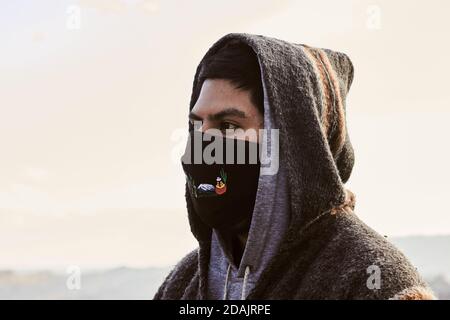 Junger hispanischer Mann mit Gesichtsmask und Kapuzenpullover, draußen während der covid-19 Pandemie Stockfoto