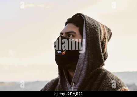 Junger hispanischer Mann mit Gesichtsmask und Kapuzenpullover, draußen während der covid-19 Pandemie Stockfoto