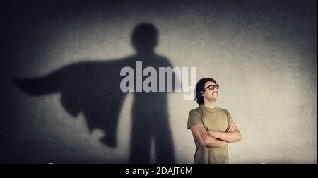 Selbstbewusster junger Mann hält Arme gekreuzt lächelnd optimistisch wie Gießen ein Superheld mit Umhang Schatten an der Wand. Motivierter und ehrgeiziger Kerl neigt zu ac Stockfoto