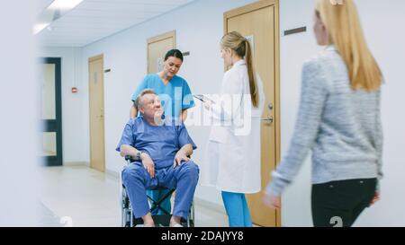 Im Krankenhausflur schiebt die Krankenschwester ältere Patienten in den Rollstuhl, der Arzt spricht mit ihnen, während er einen Tablet-Computer verwendet. Sauber, neues Krankenhaus mit Stockfoto