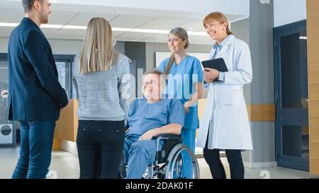 In der Krankenhauslobby. Junges Paar Besuch ältere Eltern in einem Rollstuhl, Er wird von freundlichen Krankenschwester und Arzt unterstützt. Happy Family Reunion. Neu, Modern Stockfoto
