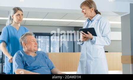 In der Krankenhauslobby schiebt die Krankenschwester ältere Patienten in den Rollstuhl, der Arzt spricht mit ihnen, während er einen Tablet-Computer verwendet. Sauber, neues Krankenhaus mit Stockfoto