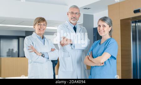 Porträt von zwei Ärzten und einer Krankenschwester, die im Krankenhausgebäude posiert, Arme gekreuzt und lächelnd. Brillante Menschen in medizinischen Profis, die sparen Stockfoto