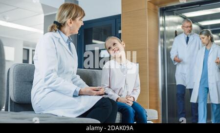 In der Lobby des Krankenhauses spricht Ärztin mit einem niedlichen kleinen Mädchen, während sie auf dem Sofa sitzt. Modernes Krankenhaus mit bestmöglicher Pädiatrie Stockfoto