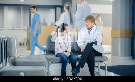 In der Lobby des Krankenhauses spricht Ärztin mit einem niedlichen kleinen Mädchen, während sie auf dem Sofa sitzt. Modernes Krankenhaus mit bestmöglicher Pädiatrie Stockfoto