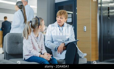 In der Lobby des Krankenhauses spricht Ärztin mit einem niedlichen kleinen Mädchen, während sie auf dem Sofa sitzt. Modernes Krankenhaus mit bestmöglicher Pädiatrie Stockfoto