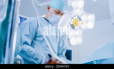 Low Angle Shot im Operationssaal, Assistentin leitet das Instrument während der invasiven Chirurgie an einen Chefarchirurgen weiter. Professionelle Ärzte, die Chirurgie in Stockfoto