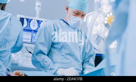 Low Angle Shot im Operationssaal, Assistentin leitet das Instrument während der invasiven Chirurgie an einen Chefarchirurgen weiter. Professionelle Ärzte, die Chirurgie in Stockfoto