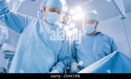 Low Angle Shot im Operationssaal von zwei Chirurgen während des chirurgischen Eingriffs Biegen über den Patienten mit Instrumenten. Professionelle Ärzte in modernen Stockfoto