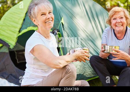 Zwei glückliche ältere Frauen sitzen vor dem Zelt Am Morgen und halten Kochtöpfe Stockfoto