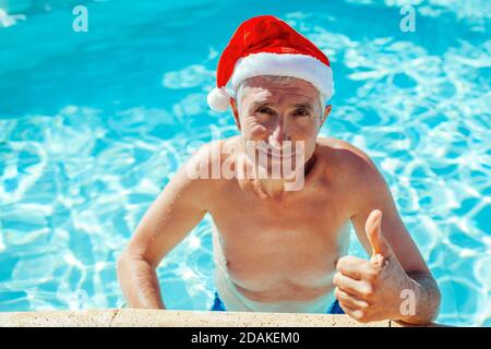 Neujahr und Weihnachtsfeier. Mann in Santa's Hut zeigt Daumen nach oben entspannen im Schwimmbad. Tropischer Urlaub Stockfoto