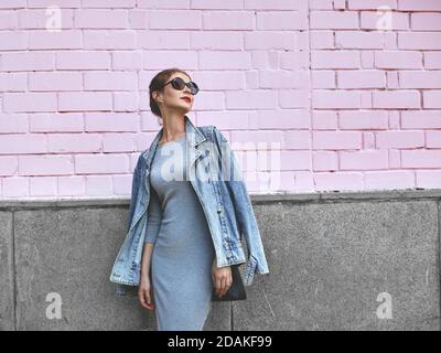 Street Style Shoot von Frau auf rosa Wand. Swag Mädchen tragen Jeans Jacke, graues Kleid, Sonnenbrille. Mode Lifestyle Im Freien Stockfoto