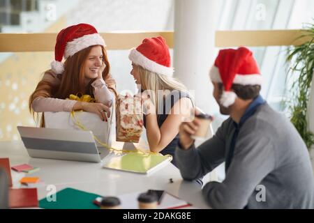 Zwei weibliche Kollegen lächeln und sprechen mit Geschenken und weihnachtsmützen im Büro, beobachtet von männlichen Angestellten Stockfoto