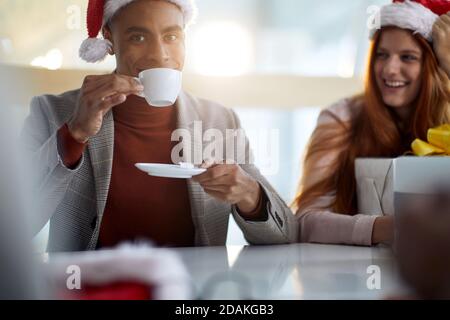 Junge afro-amerikanische Mitarbeiterin trinkt Kaffee im Büro, trägt Karo-Jacke, Rollkragenpullover und santa Hut. Blick in die Kamera Stockfoto