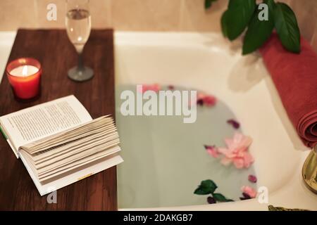 Zeit für sich selbst. Entspannen Sie sich zu Hause. Badewanne mit Blütenblättern. Buch, Kerzen und ein Glas Wein auf einem Holztablett. Bio Spa Entspannung in Komfort gemütlich Stockfoto