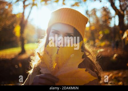Hallo oktober. Glückliche moderne 40 Jahre alte Frau in beigem Mantel und orange Hut mit Herbst gelben Blättern draußen im Stadtpark im Herbst. Stockfoto