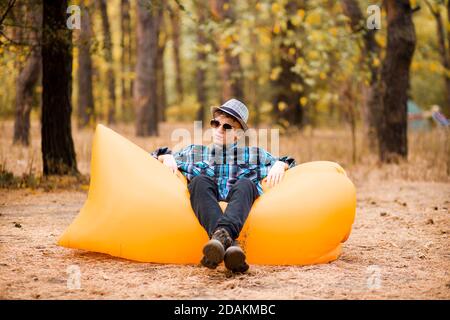 Schöner junger Mann mit Hut und Sonnenbrille auf aufblasbarem Sofa liegend. Glücklicher Mann mit Händen auf Genießen Sie freie Zeit auf der frischen Luft, während auf Gras in Fores ruhen Stockfoto