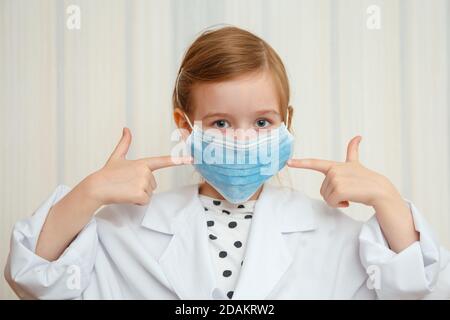 Ein kleines Mädchen im Morgenmantel und Maske eines Arztes zeigt mit einer Geste, dass jeder eine Schutzmaske tragen muss. Stockfoto