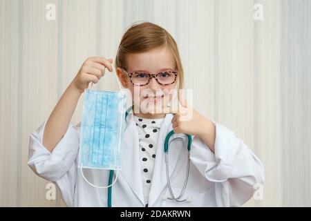 Ein kleines Mädchen im Morgenmantel und Maske eines Arztes zeigt mit einer Geste, dass jeder eine Schutzmaske tragen muss. Stockfoto