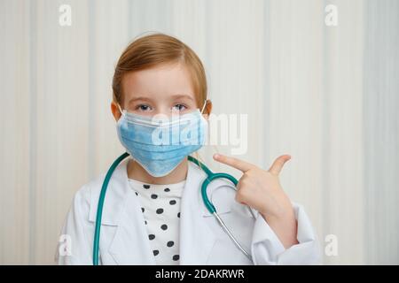 Ein kleines Mädchen im Morgenmantel und Maske eines Arztes zeigt mit einer Geste, dass jeder eine Schutzmaske tragen muss. Stockfoto