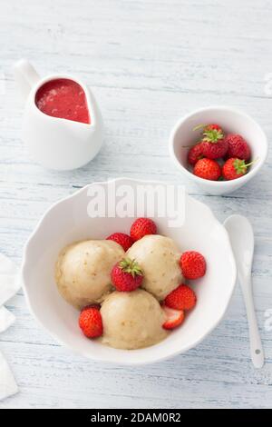 Veganes Bananeneis mit Erdbeersoße und frischen Erdbeeren In einer weißen Schale auf hellblauem Holzhintergrund Stockfoto