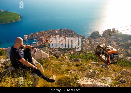 Weitaufnahme vom Gipfel des SRD-Berges, Mann auf einem Felsen sitzend, der das Gebiet um die Stadt Dubrovnik beobachtet, steigt die orangefarbene Seilbahn zum Cit hinab Stockfoto