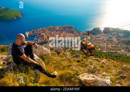 Weitaufnahme vom Gipfel des SRD-Berges, Mann auf einem Felsen sitzend, der das Gebiet um die Stadt Dubrovnik beobachtet, steigt die orangefarbene Seilbahn zum Cit hinab Stockfoto