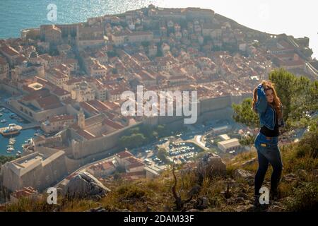 Brunette trägt blaue Jeans und blaue Spitze von hinten gesehen stehend auf dem Berg über der Stadt Dubrovnik, erstaunliche Stadt im Hintergrund wi gesehen Stockfoto