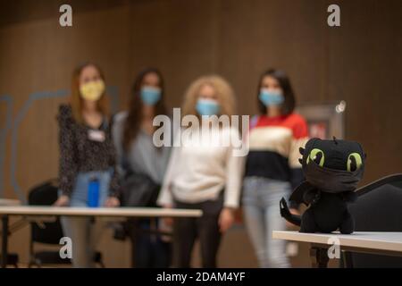 Flacher Fokus auf ein kleines Spielzeug auf dem Tisch, das eine schwarze Gesichtsmask trägt, während eine Gruppe von vier Studentinnen im Hintergrund verschwommen steht. Schüler ermüden Stockfoto