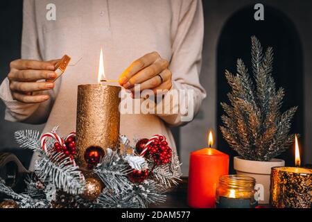 Frau Hände Blitz große goldene Kerze mit Streichhölzern in Weihnachtskranz. Weihnachten Innendekoration auf rustikalem Holzhintergrund. Hygge, Ornamente, Kerze Stockfoto