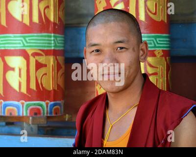 Der junge tibetische buddhistische Mönch steht vor einer Reihe von bunt bemalten großen Gebetsrädern (Manikrädern) im Tawang Kloster. Stockfoto