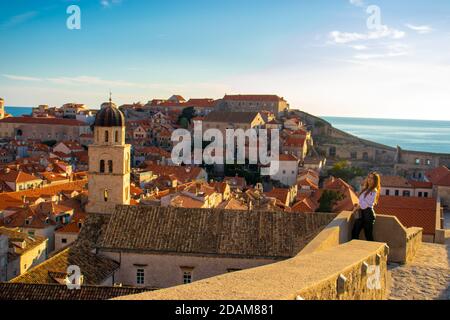 Attraktive Brünette steht auf den Stadtmauern der Altstadt von Dubrovnik, kleine Häuser und Dächer rund um die gesamte antike Gegend. Kirchenglocke Stockfoto