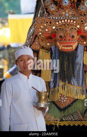 Balinesischer hinduistischer Tempelpriester in traditioneller weißer Kleidung, der neben einer Barong-Maske auf dem Gelände eines hindu-Tempels im Zentrum von Bali, Indonesien, steht Stockfoto