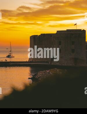 Goldener Sonnenuntergang in der Stadt Dubrovnik, Schloss am Ufer eines kleinen Hafens, Segelboot verlässt den Hafen und ein kleines lokales Fischerboot e Stockfoto