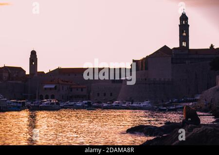 Schöner Sonnenuntergang über der Altstadt von Dubrovnik. Kleiner lokaler Hafen vor der Stadt, Frau in schwarzen Kleidern sitzt auf einem nahe gelegenen Felsen und beobachtet t Stockfoto
