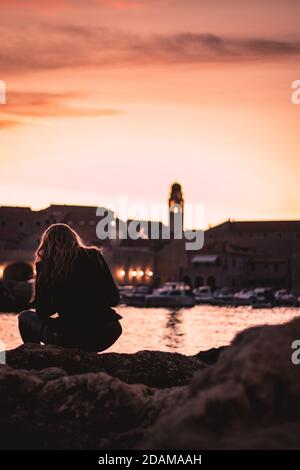 Schöner Sonnenuntergang über der Altstadt von Dubrovnik. Kleiner lokaler Hafen vor der Stadt, Frau in schwarzen Kleidern sitzt auf einem nahe gelegenen Felsen und beobachtet t Stockfoto
