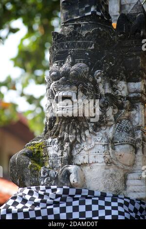 Rakasa - mythischer Hindu-Dämon-Wächter mit langen Zähnen am Tempeleingang, Bali, Indonesien. Mit schwarz-weiß kariertem Polniertuch verziert Stockfoto