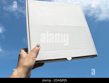 Eine Frauenhand hält eine weiße Pappschachtel mit einer Kopie des Raumes und des Platzes für Text auf einem blauen Himmel Hintergrund, Schablone, mockup Stockfoto