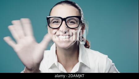 Fröhliche junge Frau, die die Kamera anschaut und mit ihr winkt Hand Stockfoto