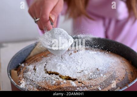 Frau Hand bestreuen Puderzucker über frischen Biskuitkuchen. Puderzucker fällt auf frischen perfekten Marmorkuchen/ Stockfoto
