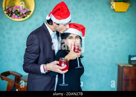 Junges Paar in santa Hüte Weihnachten feiern zusammen halten roten Cocktail trinken Gläser Freund küsst auf ihre Freundinnen Stirn, Liebe, bondi Stockfoto