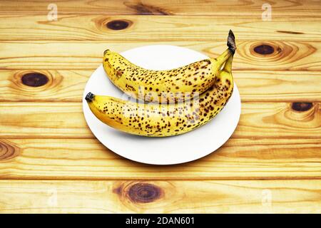 Reife gesprenkelte Bananen auf einem weißen Teller. Draufsicht, Holztisch Hintergrund. Stockfoto