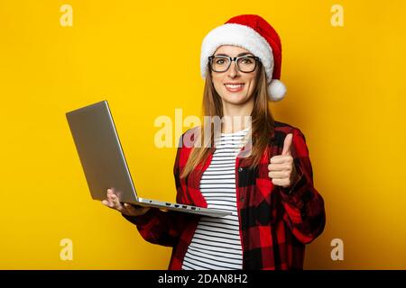 Junge Frau in einem weihnachtsmütze und einem roten Hemd in einem Käfig hält einen Laptop in den Händen und macht eine Geste alles ist gut auf einem gelben Hintergrund. Stockfoto