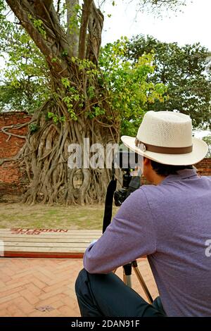 Mann in weißem Hut Bilder des berühmten Kopf der alten Buddha-Statue gefangen in Bodhi Tree Roots in Wat Mahathat Temple Ruins, Ayutthaya, Thailand Stockfoto