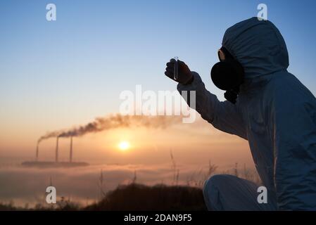 Nahaufnahme eines Ökologen in einer Gasmaske, die das Reagenzglas mit der Bodenprobe hält. Wissenschaftler, die Umweltprobleme im Kraftwerk mit rauchenden Kaminen und schönem Himmel im Hintergrund erforschen. Stockfoto